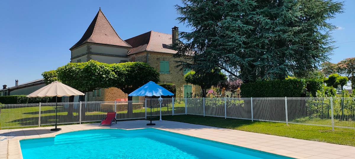 A spacious outdoor area is displayed, featuring a secure swimming pool with clear blue water. Surrounding the pool are shaded lounging areas with umbrellas. The elegant house is set against a backdrop of well-maintained greenery, offering a tranquil atmosphere.