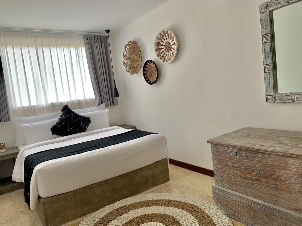 A bright bedroom features a spacious bed with crisp white linens and a dark accent blanket. Decorative wall art and a round woven rug complement the light-colored walls. A wooden storage chest and a mirror are positioned adjacent to the bed, enhancing the room's functionality.