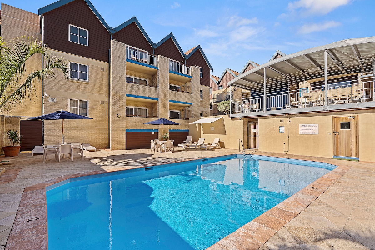 A communal pool area is featured, surrounded by lounge chairs and umbrellas. The pool's clear blue water reflects the sunny sky above. Nearby, a covered seating area is visible, providing shade for guests. The building's facade and balconies offer additional charm to the outdoor space.