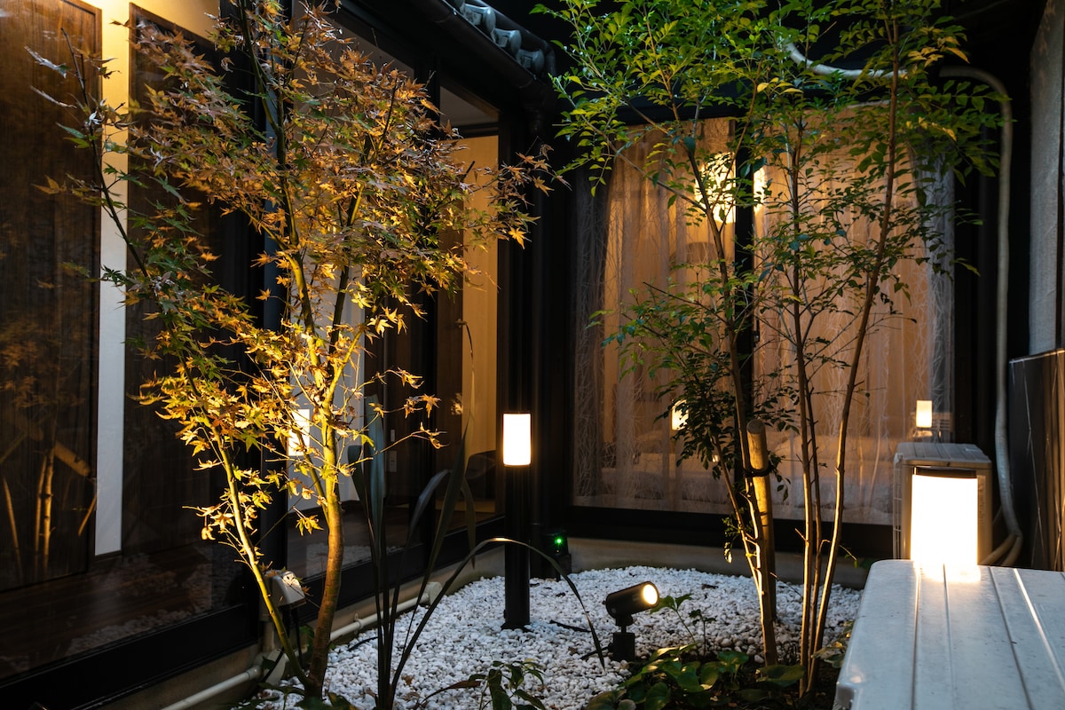 An atmospheric courtyard is illuminated by soft outdoor lights, featuring thoughtfully arranged trees and subtle landscaping with white stones. The surrounding glass walls create a serene blend of indoor and outdoor spaces, enhancing the tranquil ambiance at night.