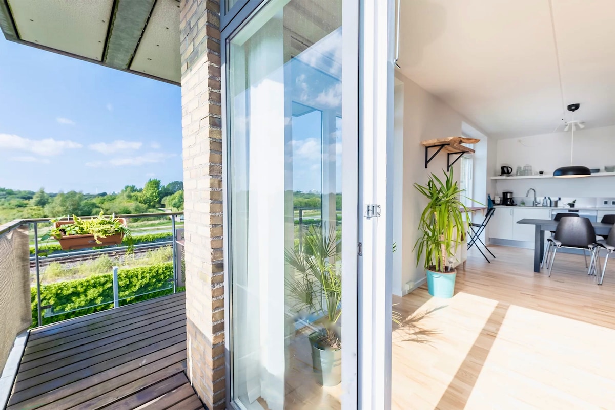 A bright corner of the apartment reveals a glass door that opens to a spacious balcony. Potted plants adorn the balcony, while an open layout showcases the living area with modern furnishings and a well-equipped kitchen in the background.