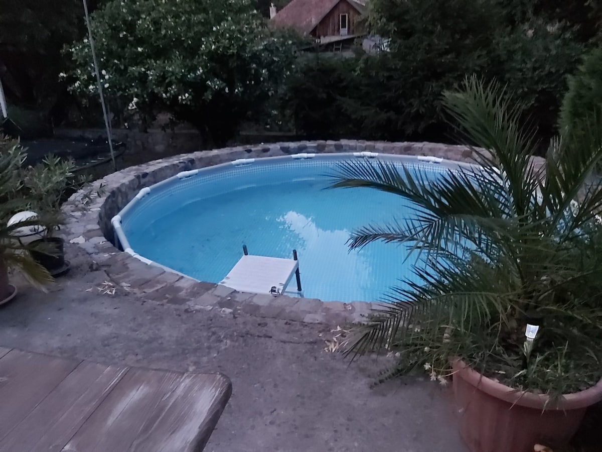 A circular pool is surrounded by a stone border, with clear blue water reflecting the evening sky. Lush green foliage is visible nearby, enhancing the natural setting. A wooden platform is situated at one edge of the pool, providing access.