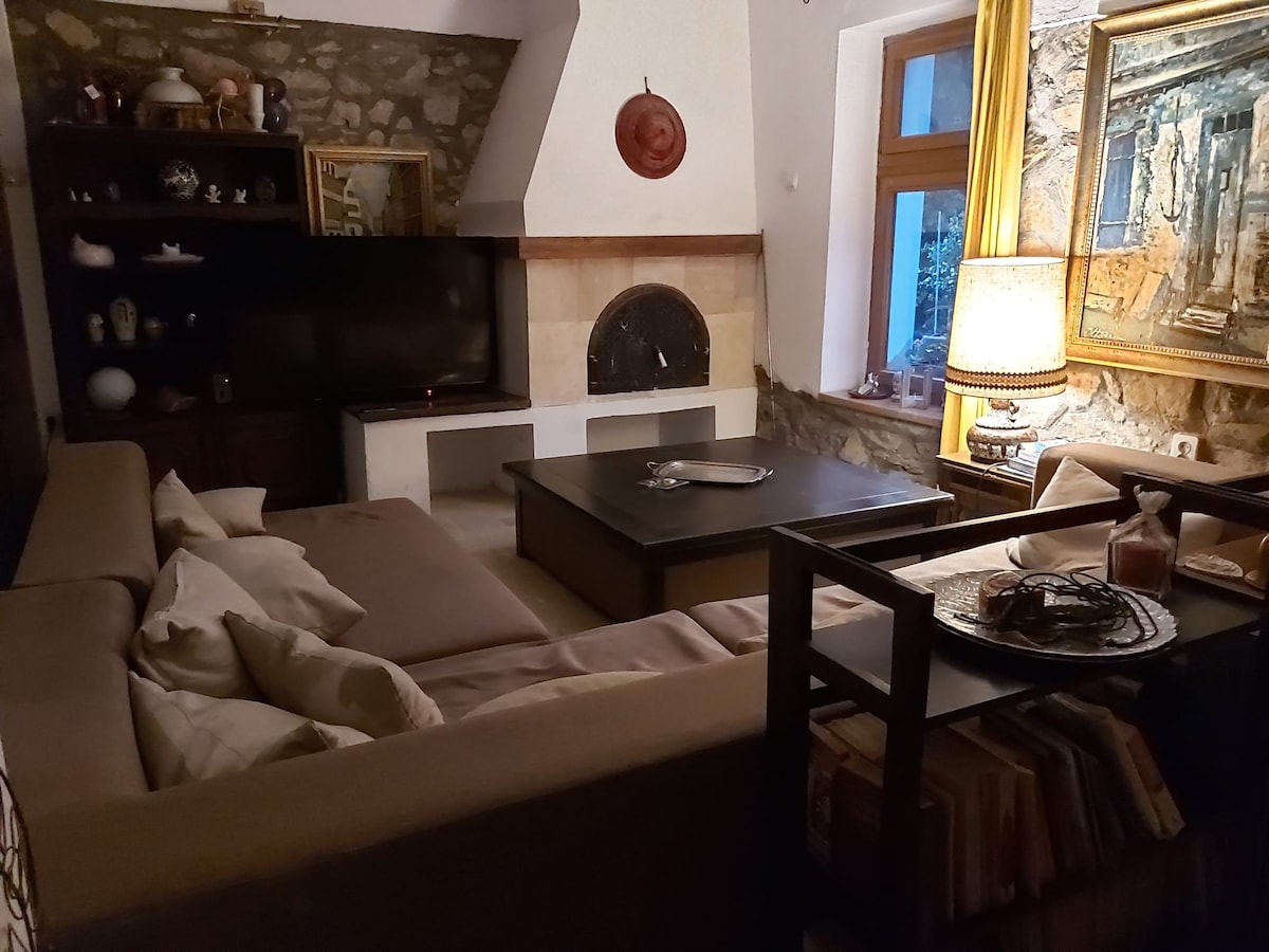 A spacious living area features a large, L-shaped sectional sofa with light-colored cushions. A dark coffee table sits in the center, complemented by decorative items. A flat-screen television is positioned against a stone wall, while warm light emanates from a nearby lamp, illuminating a cozy ambiance.