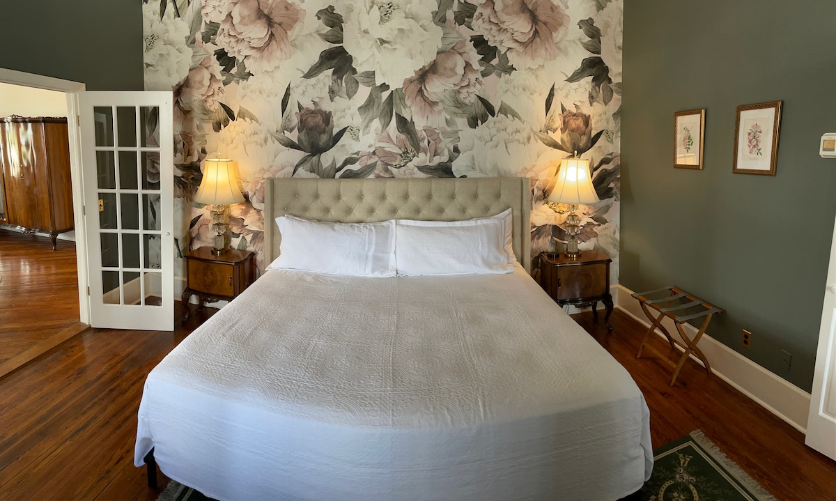 A spacious sleeping area is highlighted by a plush bed with white bedding, set against a floral accent wall adorned with large peonies. Two bedside lamps provide soft lighting, and light-colored wooden furniture complements the warm hardwood floors.