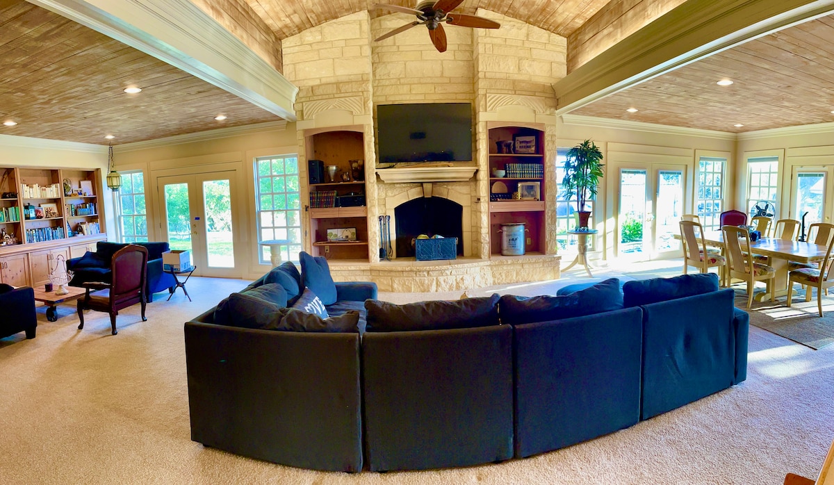 A spacious living area showcases a large sectional sofa facing a stone fireplace with a mounted television above. Natural light floods in through multiple windows, illuminating built-in shelves filled with books. A dining table can be seen in the background, enhancing the open layout.