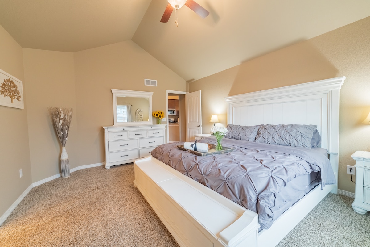 A spacious bedroom features a large bed with a decorative gray coverlet and two bedside lamps. A dresser and mirror are positioned against the wall. The room is softly illuminated, highlighting the vaulted ceiling and neutral wall color, creating a calming atmosphere.