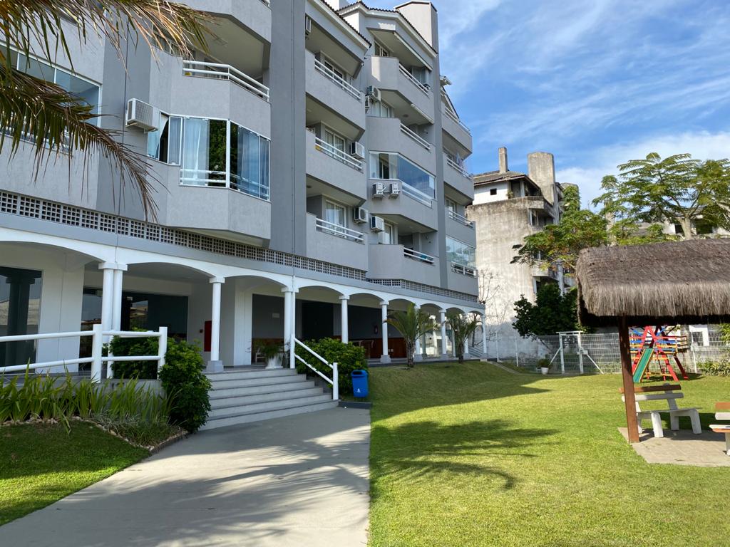 The exterior of a multi-story building features a light-colored facade with balconies and large windows. A lawn area is visible in front, with well-maintained greenery and a playground in the background. A thatched-roof structure adds an inviting touch to the outdoor space.