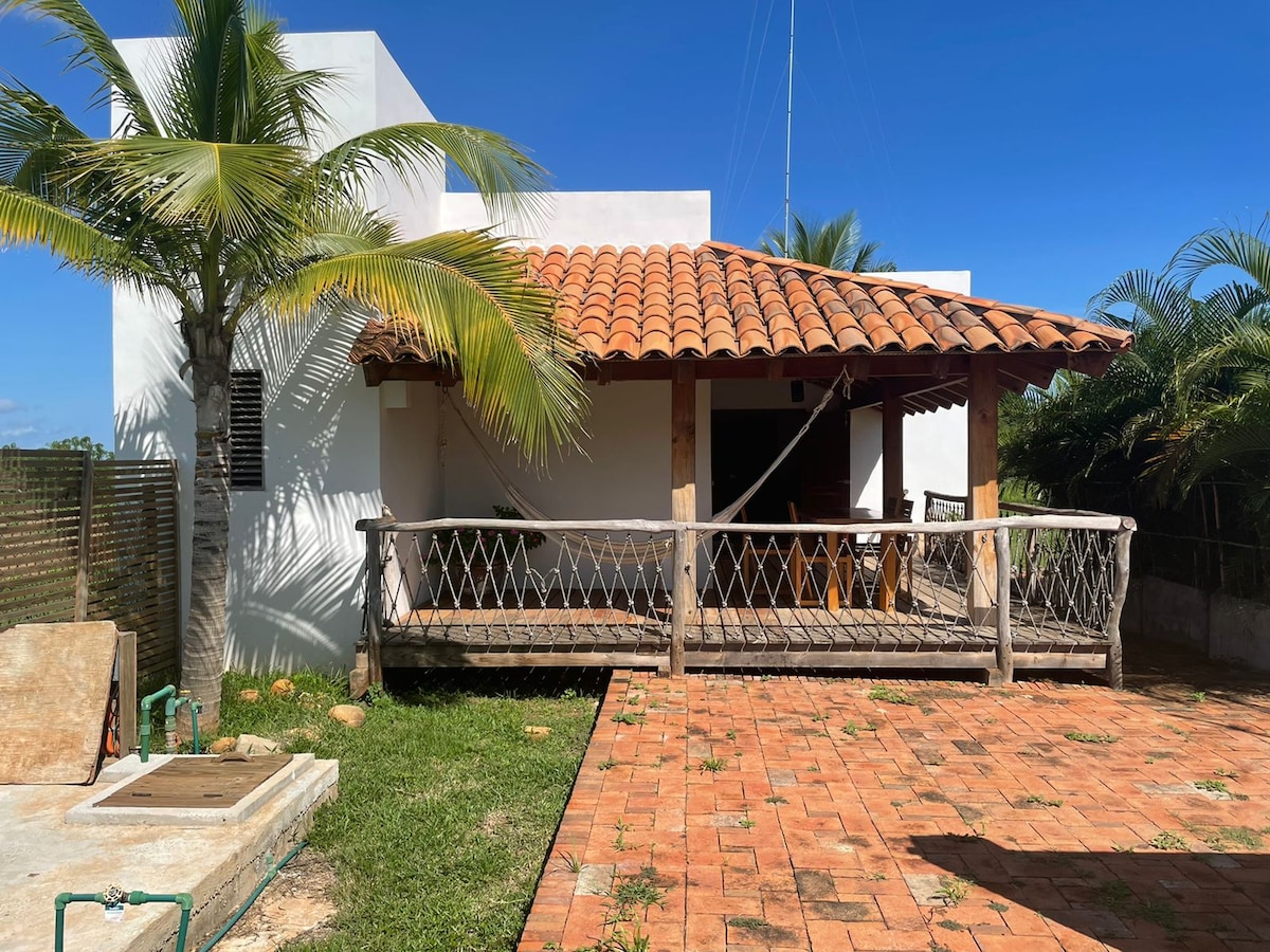 A welcoming outdoor deck is framed by a sturdy wooden railing, featuring a hammock for relaxation. The structure showcases a traditional tiled roof and white walls. Vibrant palm trees surround the space, with terracotta tiles extending across the ground, inviting moments of leisure in a tranquil setting.