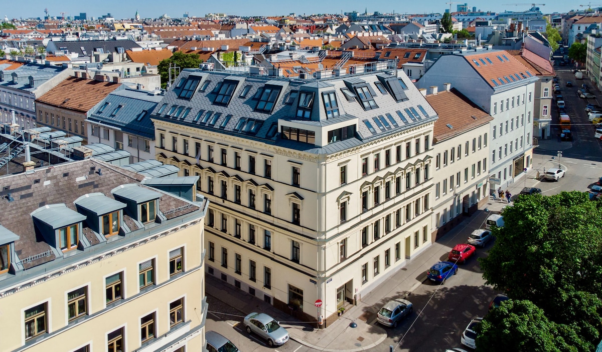 The apartment building showcases a blend of classic and modern architecture, featuring a light-colored facade and large rooftop windows. Surrounded by a variety of residential and commercial structures, the scene offers a vibrant urban atmosphere with elements of greenery included.