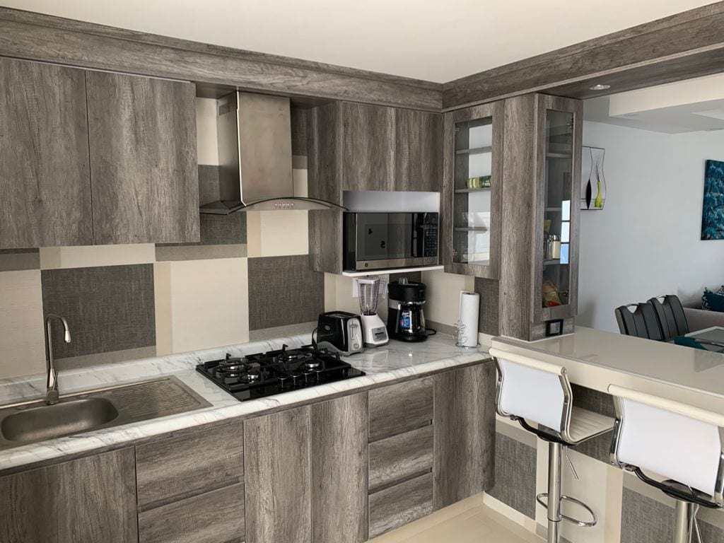 A modern kitchen is presented with a blend of wood and stone finishes. Equipped with essential appliances, including a gas stove, microwave, and coffee maker, it features a sleek countertop and two high stools. Ample cabinetry is available for storage.