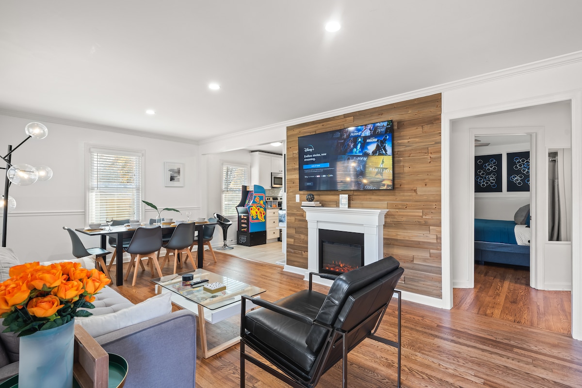 A comfortable living area is highlighted by a modern black chair and a sleek coffee table, surrounded by wood accents. A wall-mounted TV displays streaming options beside a cozy electric fireplace. The open layout leads towards a dining area and kitchen, enhancing the spacious atmosphere.