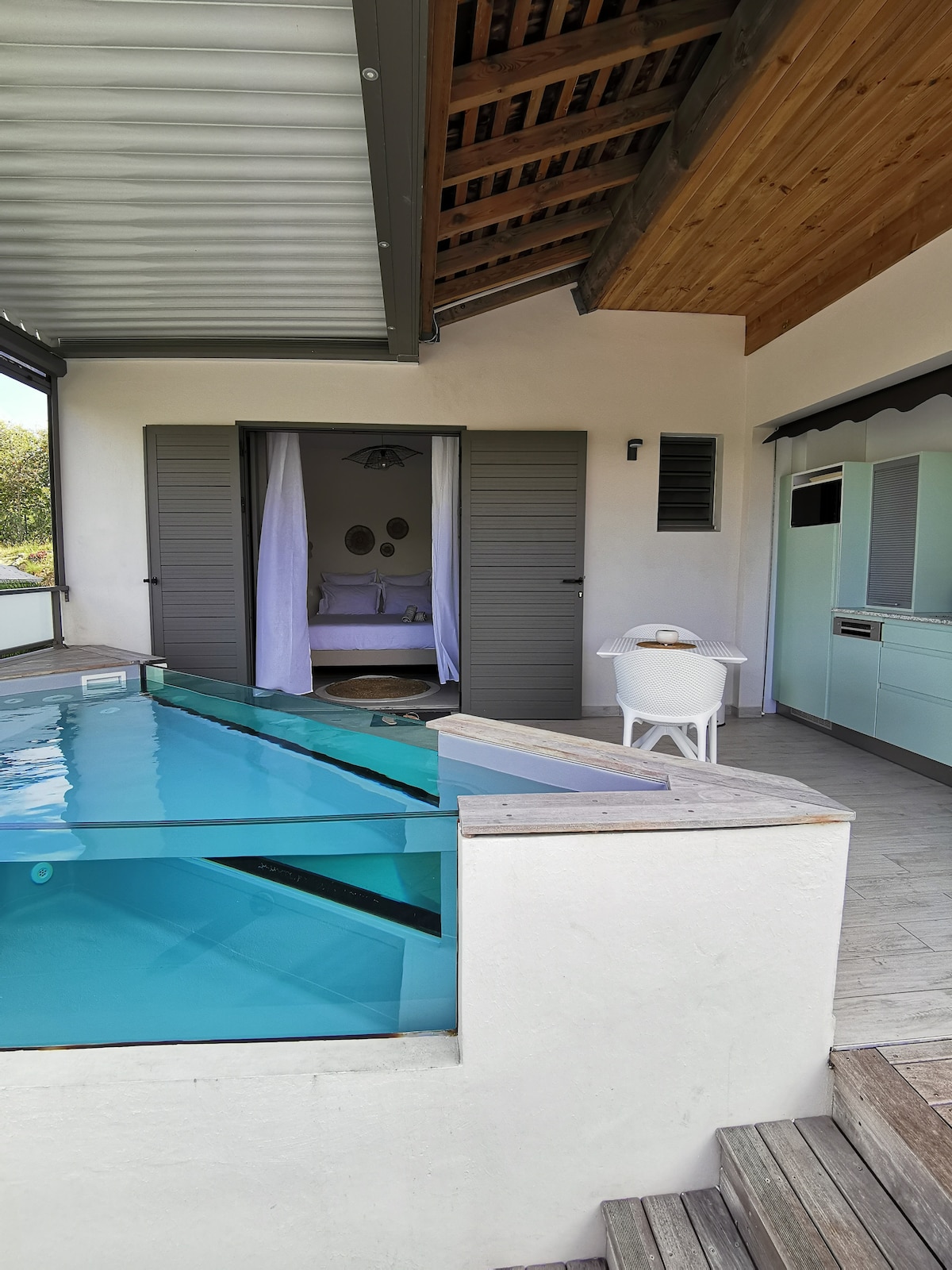 A modern outdoor space features a triangular private pool with clear water. The pool is bordered by a wooden deck leading to sliding glass doors, which open to a serene sleeping area beyond. A small dining table and chairs are positioned nearby.