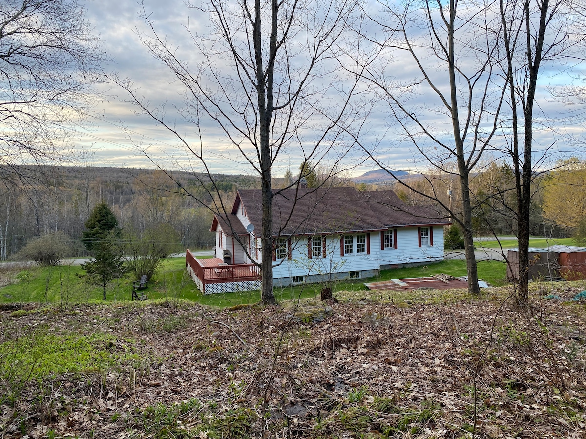 A scenic view of the cabin is captured from a distance, showcasing its white exterior with red accents. Surrounded by trees, the cabin features a spacious deck, while a gentle slope leads to an open green area and distant hills under an overcast sky.