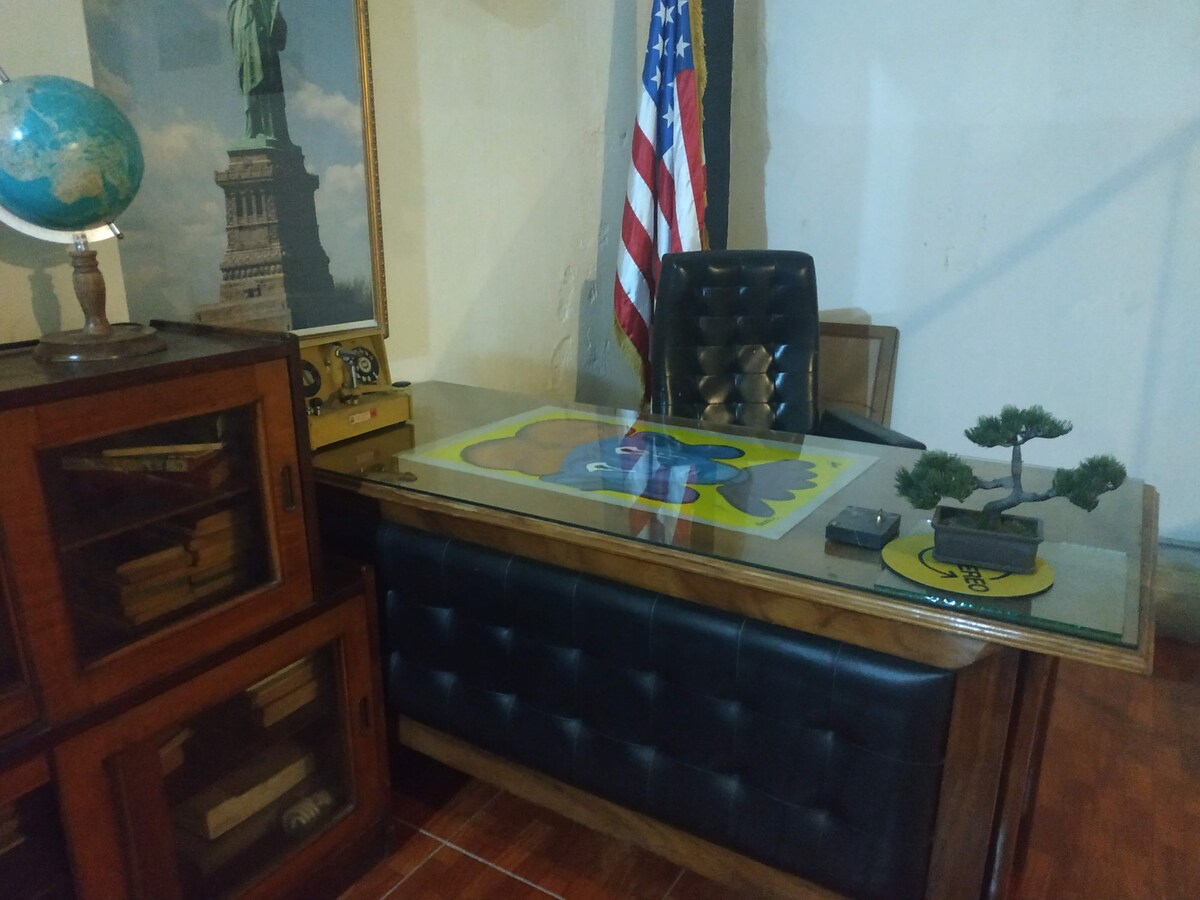 An office space is portrayed, featuring a large wooden desk with a glass top. A black leather office chair is positioned behind it. A small potted bonsai tree and a globe are displayed on the desk. The American flag and a framed image of the Statue of Liberty are visible on the wall.