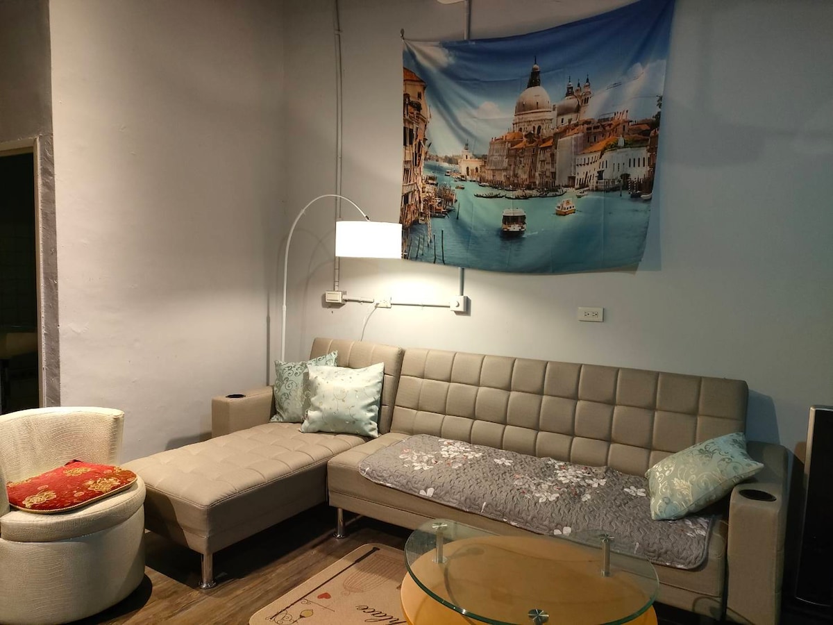 A spacious living area features a sectional sofa and an armchair, complemented by a circular glass coffee table. A cityscape tapestry hangs on the wall, adding visual interest. Soft lighting is provided by a floor lamp, creating a comfortable ambiance.