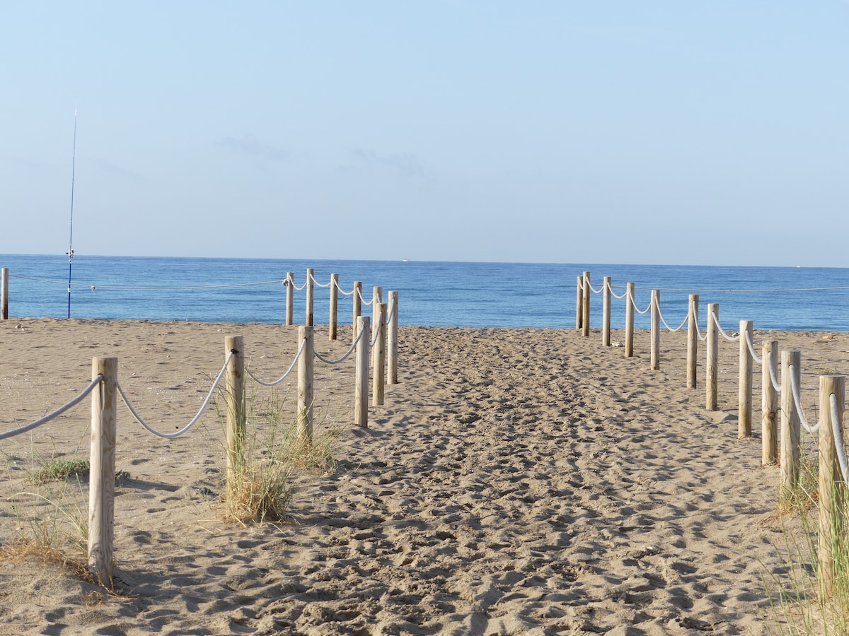 A sandy path leads to the tranquil waters of the sea, framed by wooden posts with rope rails. The beach is open and spacious, with gentle waves visible in the distance under a clear sky.