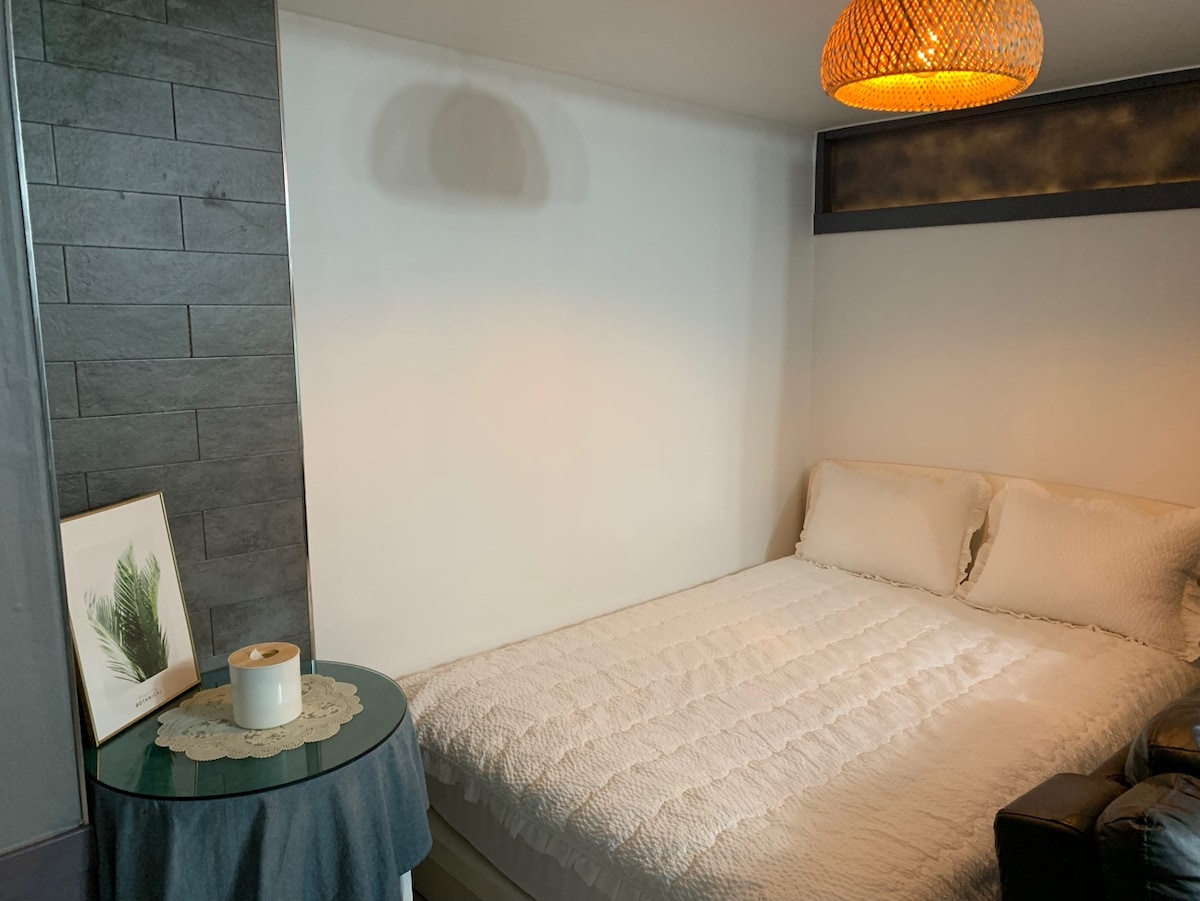 A cozy sleeping area features a spacious bed dressed in a white quilt. Next to the bed, a small round table is adorned with a decorative cloth and a candle. A basket-like light fixture casts a warm glow across the room.