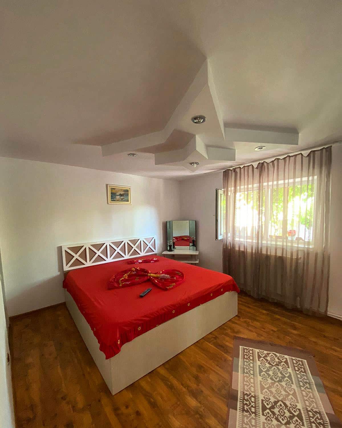 A bedroom features a spacious bed adorned with a red bedspread. Natural light enters through a window, which is dressed with sheer curtains. A full-length mirror is positioned nearby against a light wall, complemented by a patterned rug on the hardwood floor.