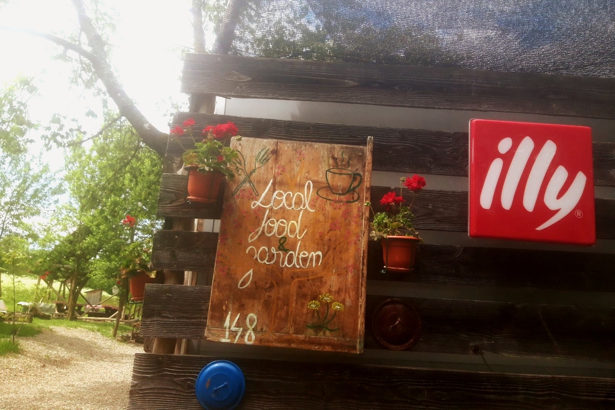 A wooden sign displaying 'Local Food Garden 148' is mounted on a rustic wall, surrounded by potted red flowers. A logo for Illy coffee is visible on the adjacent wall. Natural greenery and pathways are seen in the background, suggesting a welcoming outdoor ambiance.