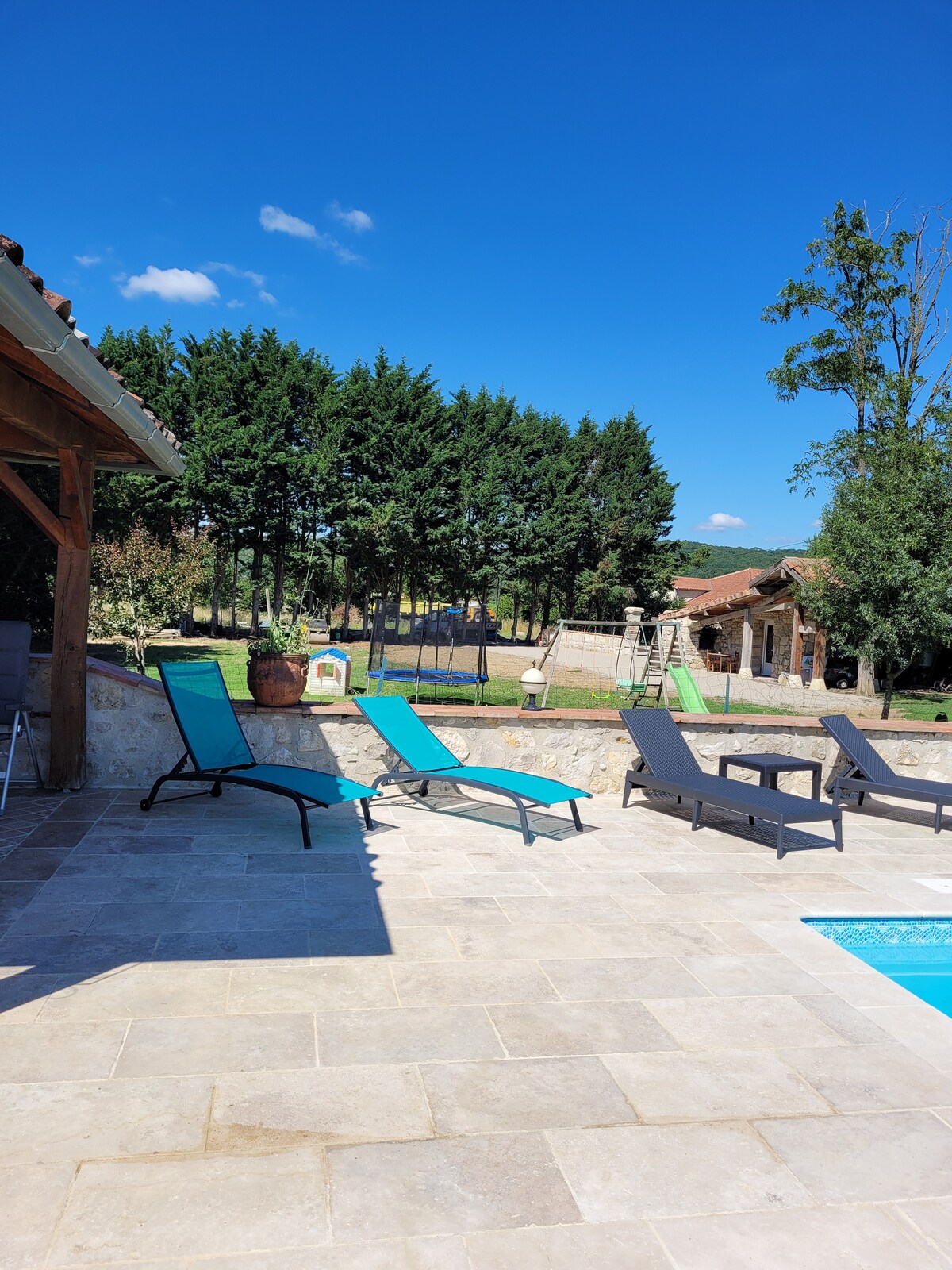 A sunlit outdoor area is present, featuring lounge chairs arranged on a stone patio beside a swimming pool. Lush greenery lines the background, with a play structure visible. The clear blue sky enhances the tranquil atmosphere of the setting.