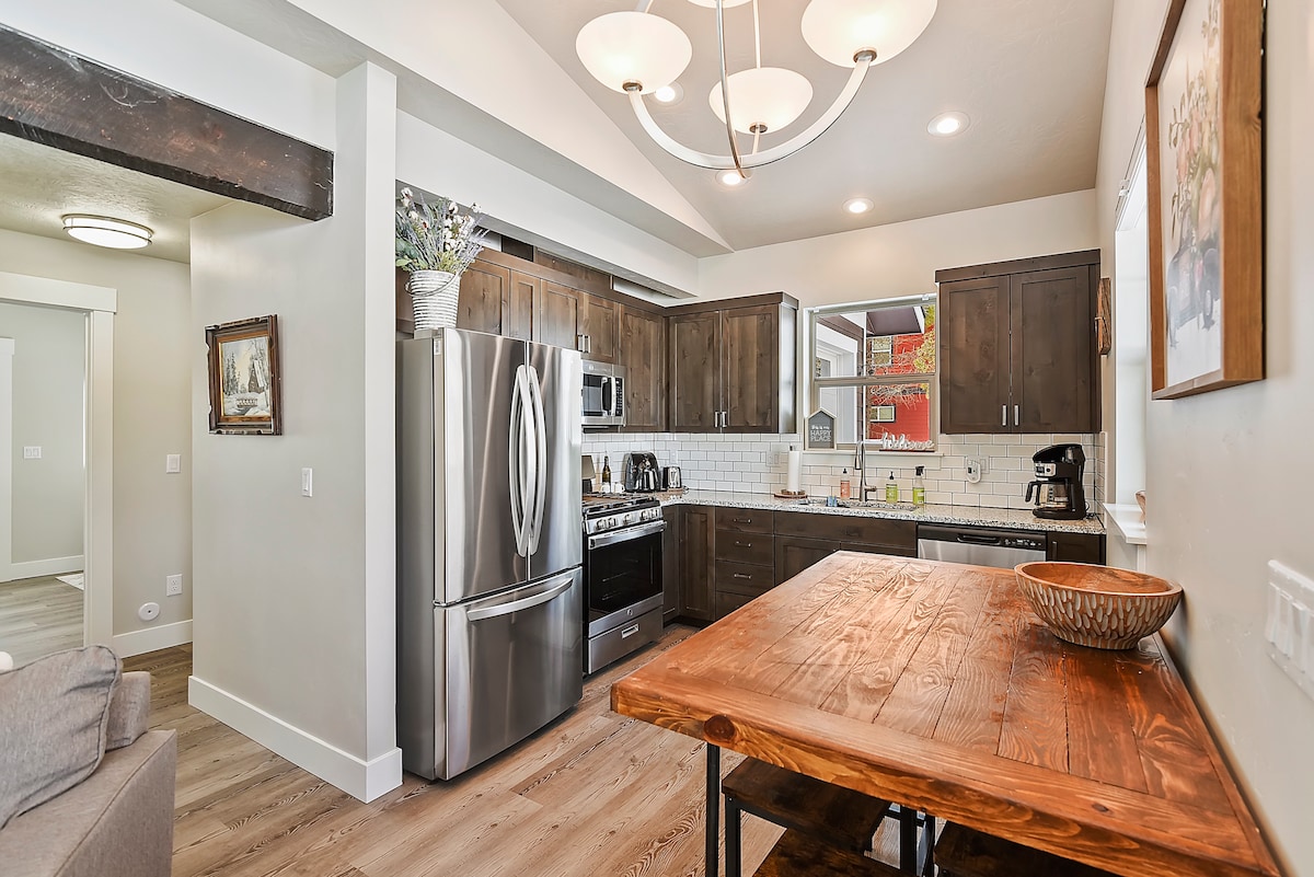 The kitchen showcases dark wood cabinetry and stainless steel appliances, including a refrigerator and stove. A wooden dining table is positioned nearby, enhancing the inviting atmosphere. Natural light pours in through windows, highlighting the clean and functional layout.