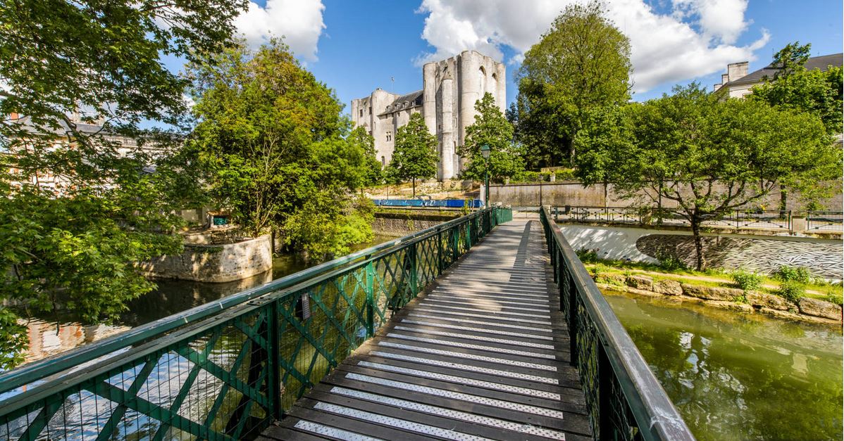 A green metal bridge leads across a serene waterway, with lush trees lining both sides. In the background, a historic castle stands amid vibrant greenery under a partly cloudy sky, creating a peaceful outdoor setting.