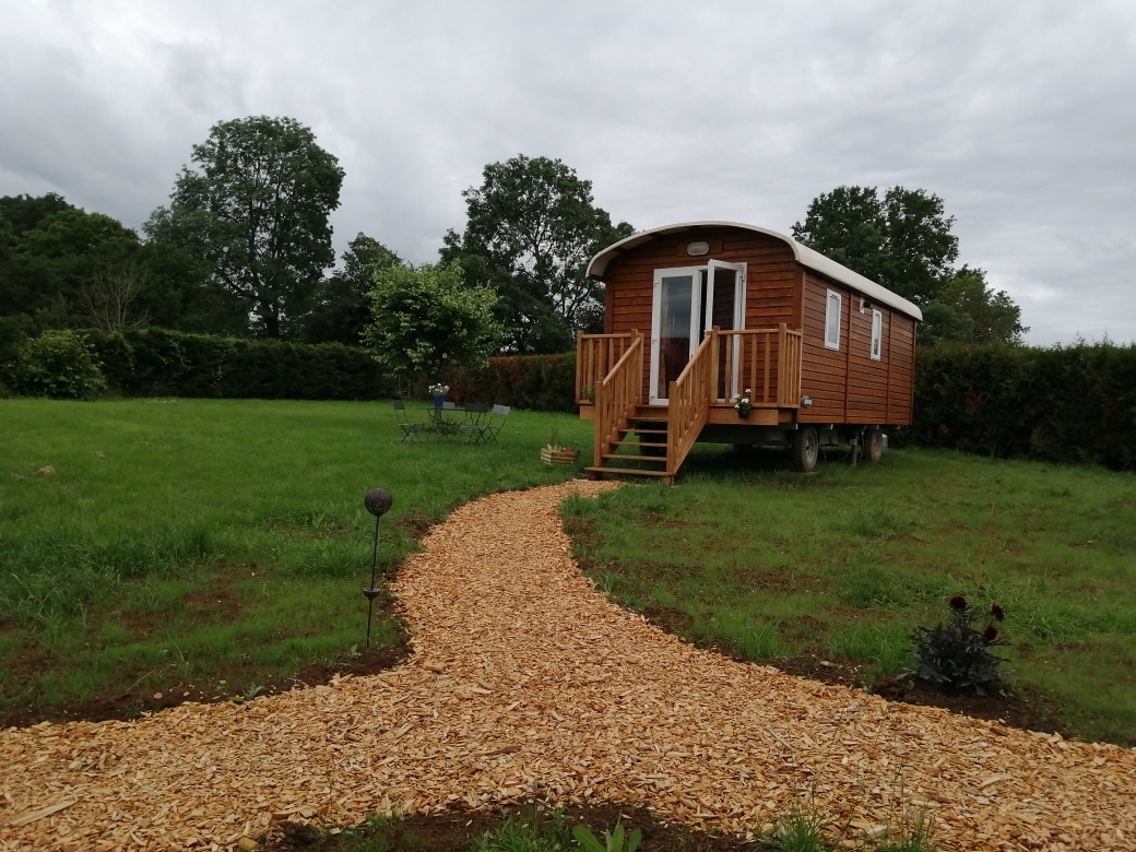 A charming wooden caravan rests on a spacious grassy area, surrounded by greenery. A walking path made of wood chips leads to the entrance, inviting visitors to explore. The exterior features large windows and a welcoming front porch, enhancing the rustic appeal.