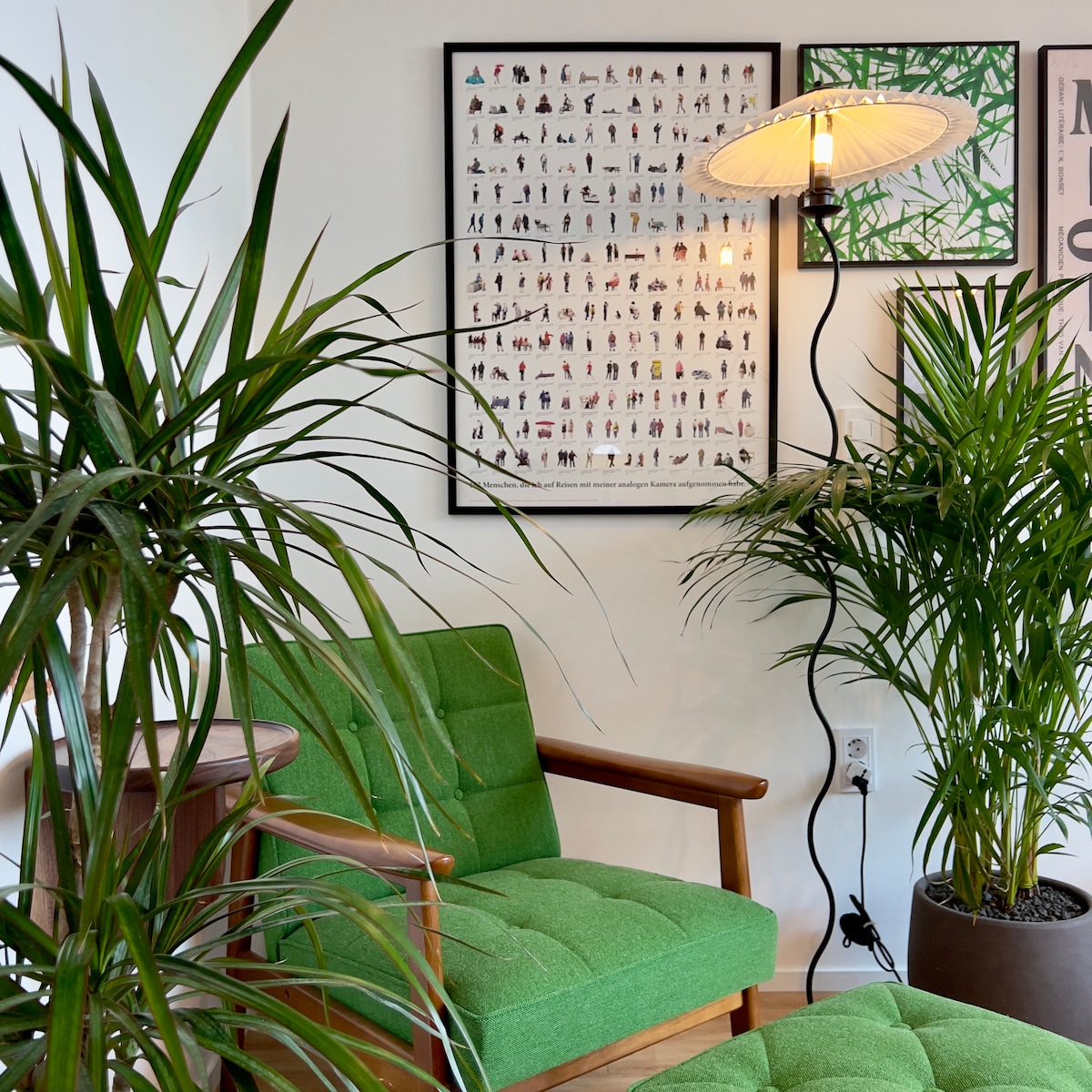 A green cushioned armchair is positioned beside a tall potted plant, adding a natural touch to the room. Framed art featuring diverse figures decorates the wall, and a floor lamp with a round shade provides soft illumination, enhancing the welcoming atmosphere.
