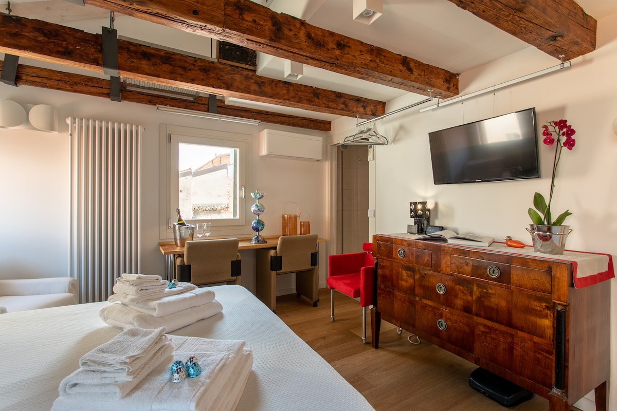 The room features rustic wooden beams overhead, complemented by modern furnishings. A spacious bed is adorned with neatly arranged linens. A wooden dresser and a small workstation with chairs provide functional space. Natural light fills the room through a window, enhancing the inviting atmosphere.