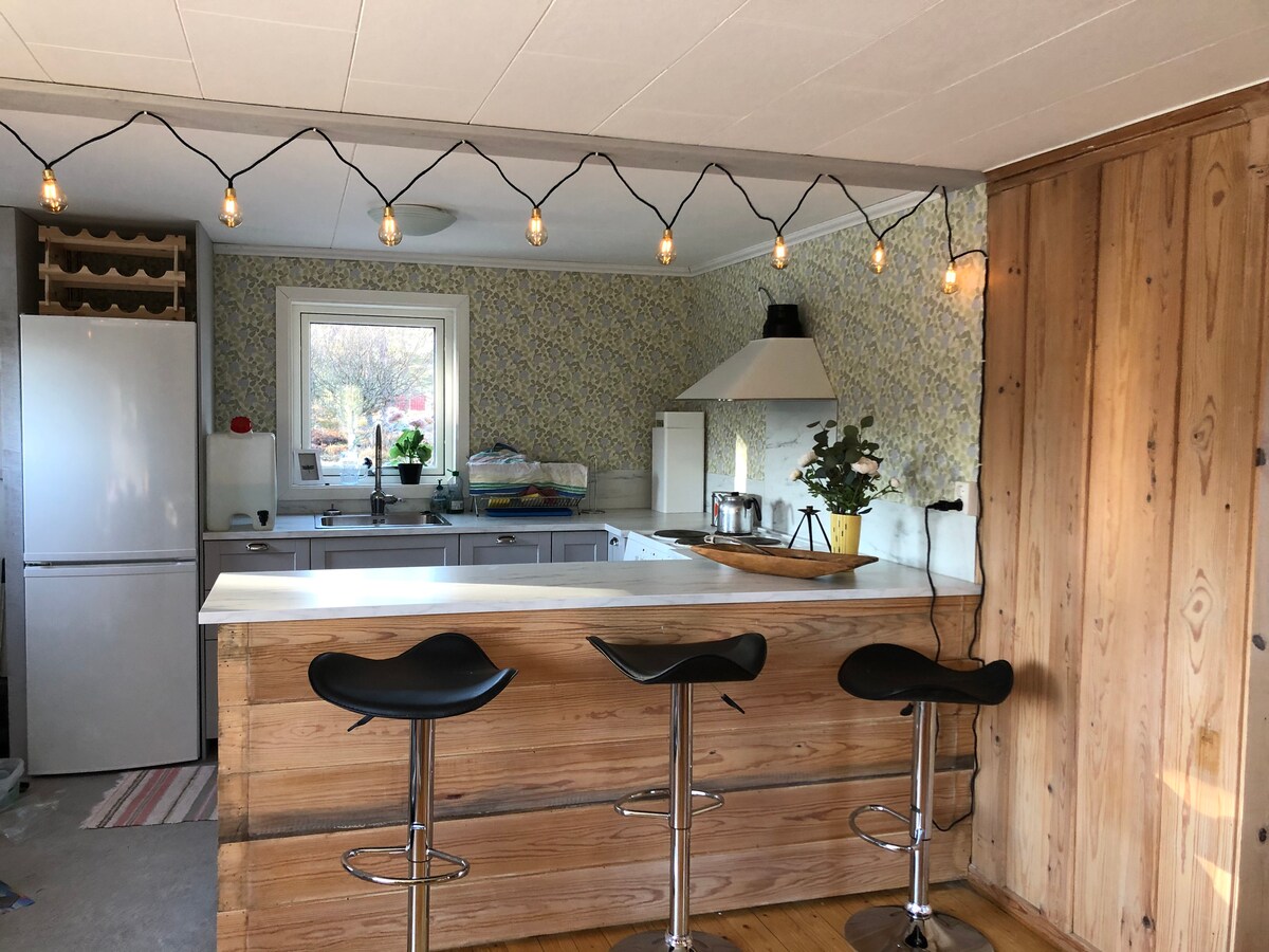 A newly renovated kitchen area features a large white countertop with three black bar stools. Soft lighting hangs above, complementing the wood accents and patterned wallpaper. Appliances include a refrigerator and a stove, with a window offering natural light and a view of the outdoors.