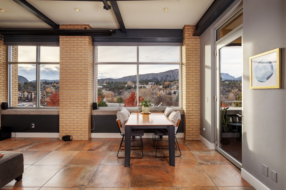A bright dining area features a wooden table surrounded by four chairs and is illuminated by natural light from expansive windows. Scenic mountain views can be seen through the glass panes, with hints of colorful foliage outside. A door leads to an outdoor space.