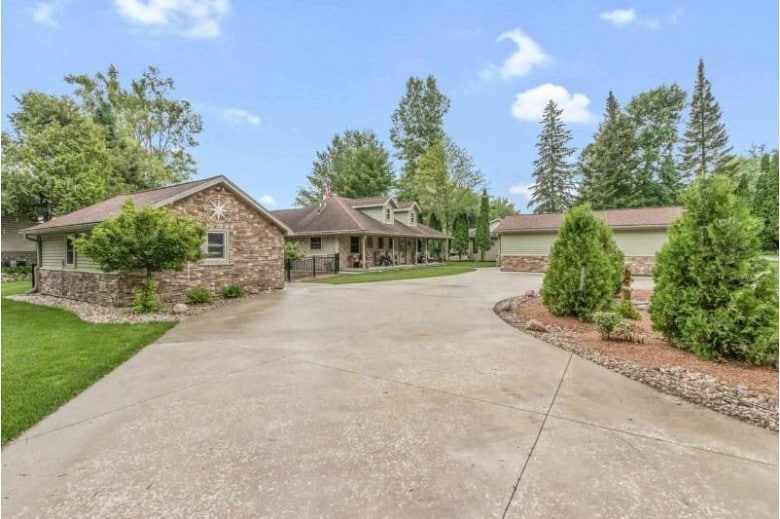 A spacious circular driveway is showcased, providing ample parking for multiple vehicles. Lush greenery surrounds the property, with neatly trimmed shrubs and trees enhancing the natural setting. The single-story home displays a stone facade, blending harmoniously with the landscaped outdoor area.
