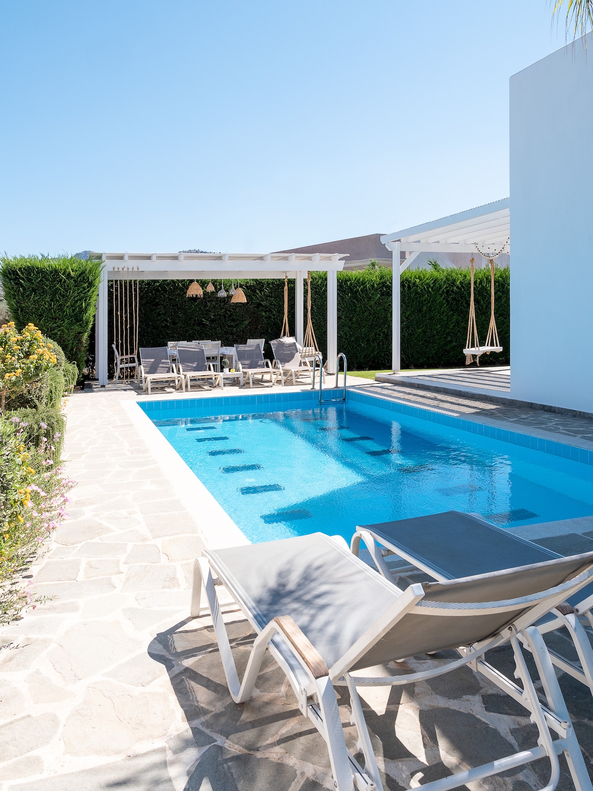 A serene outdoor space is highlighted by a private pool complemented by lounge chairs. A nearby pergola provides shaded seating, with swing chairs adding to the relaxing environment. Lush greenery surrounds the area, contributing to the inviting atmosphere of leisure and comfort.