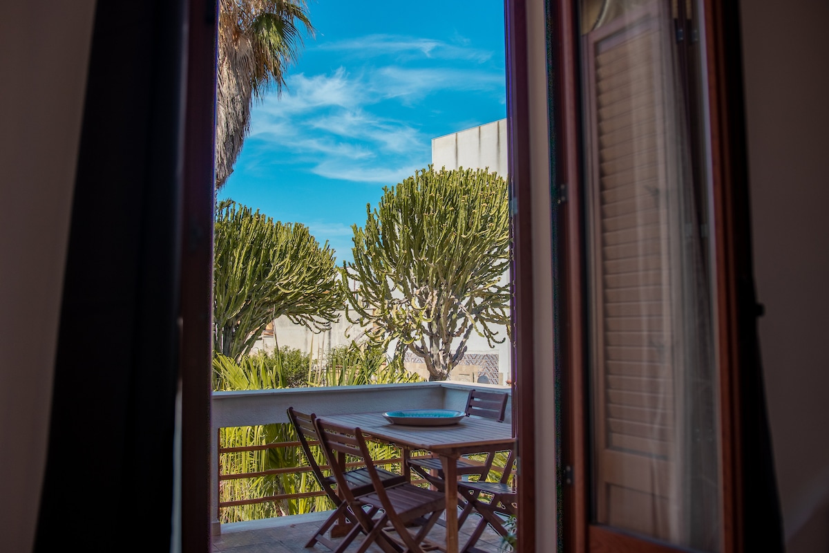 A view is captured from an open window, showcasing a balcony with a wooden table and chairs. Lush greenery, including tall plants, is visible in the background against a clear blue sky.