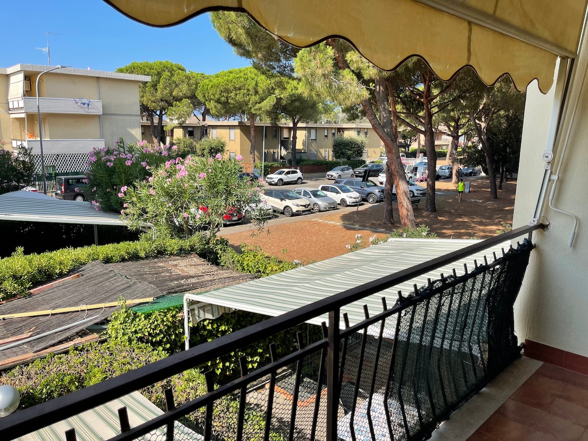 A covered terrace view showcases a peaceful courtyard with adjacent parking spaces. Lush greenery and blooming flowers line the perimeter, while nearby buildings provide a backdrop. The scene is brightened by clear skies, and a protective awning overhangs the terrace railing.