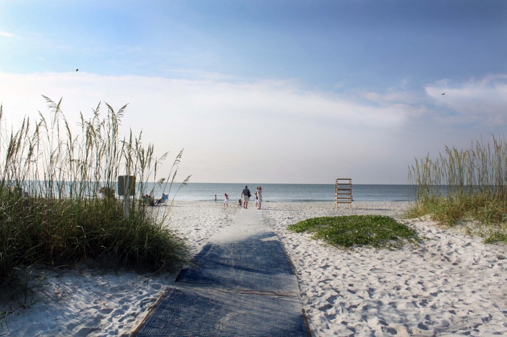 A sandy beach path leads through gentle dunes of sea oats and grass toward the tranquil sea. The calm water reflects the sky under soft clouds, while a few beachgoers can be seen enjoying the shoreline, with a lifeguard chair positioned prominently in the background.