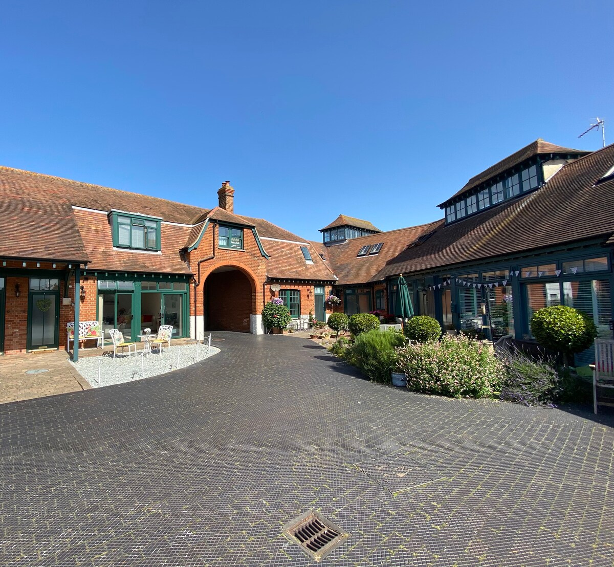 The entrance to the mews house is shown, featuring a spacious courtyard surrounded by lush greenery and well-maintained shrubs. Outdoor seating is visible, inviting guests to enjoy the tranquil surroundings under a clear blue sky.