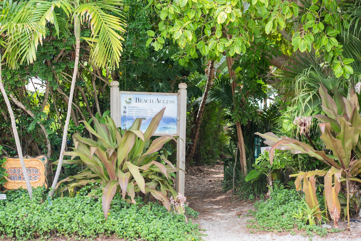An inviting pathway leads through lush greenery, framed by tropical plants on either side. A sign indicating beach access is visible at the entrance, guiding guests to the nearby beach. Soft sunlight filters through the leaves, enhancing the serene environment.