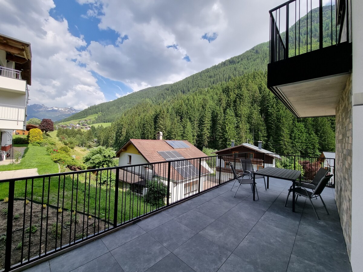 An expansive terrace is featured, framed by a wrought iron railing. A dining table and chairs are positioned for outdoor enjoyment. Lush green hills and scattered woods are visible in the background, under a partly cloudy sky.