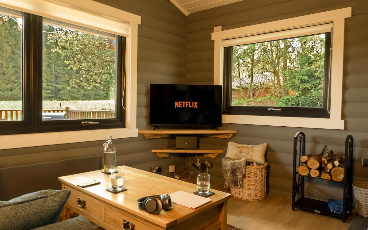 A cozy living area is presented with large windows framing the surrounding greenery. A television screen displays the Netflix logo. A wooden coffee table is accompanied by a water pitcher and glasses, while a basket holds firewood nearby, adding a homey touch.