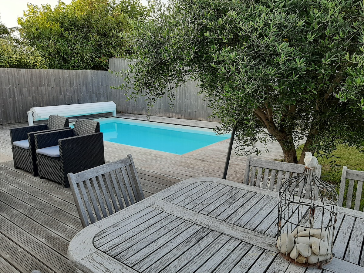 An outdoor space features a rectangular pool surrounded by a wooden deck. A dining table with wooden chairs is positioned nearby, complemented by a decorative birdcage. In the background, lush greenery and a wooden fence enhance the serene environment.
