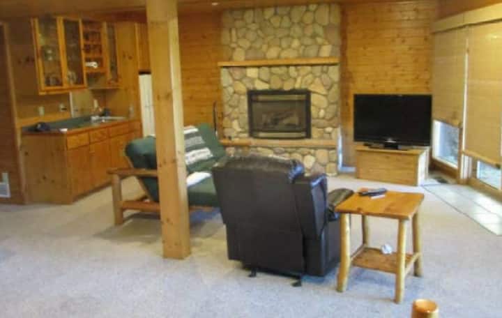 Pohľad na nižšiu obývaciu izbu s plynovým krbom a jednou z dvoch postelí/pohoviek v štýle futon.  Mokrý bar/kuchynský kút s 2. chladničkou vľavo.  Spálňa na nižšej úrovni bezprostredne po chodbe vľavo, hneď za výhľadom. 