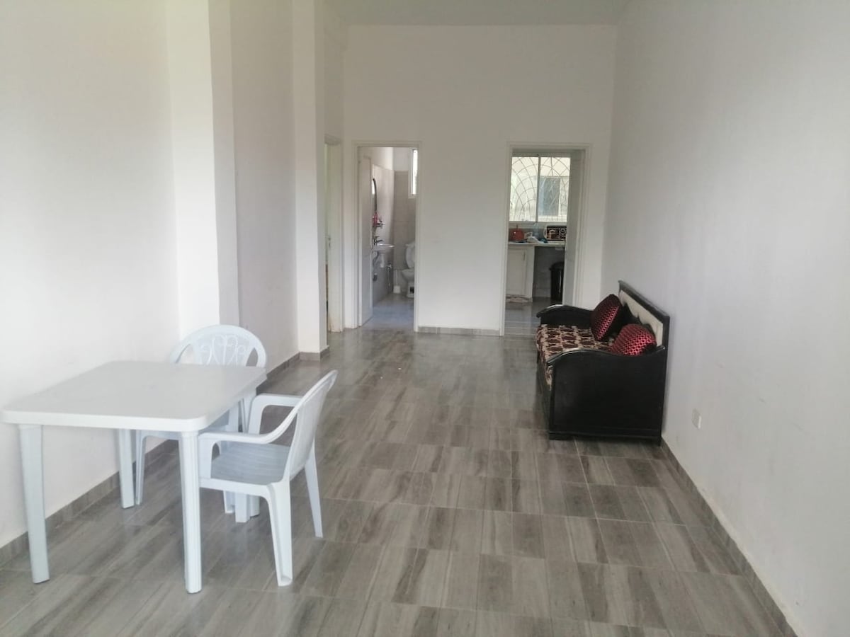 An open living area features light-colored walls and a tiled floor. A small dining table with four chairs is positioned near the window, while a black couch with decorative pillows stands against the wall, creating a simple and inviting space.