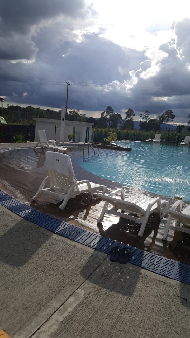 A serene outdoor pool is surrounded by lounge chairs, inviting relaxation. The water reflects the cloudy sky, creating a peaceful atmosphere. Lush greenery is visible in the background, enhancing the tranquil setting.