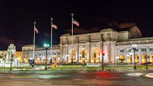 The exterior of Union Station is depicted at night, showcasing its grand architecture with large arched windows and detailed stonework. Five flags are seen waving in the breeze, illuminated by streetlights that reflect off the nearby pavement.