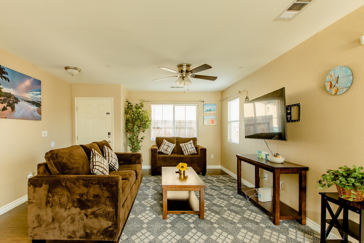 The living room features a comfortable seating arrangement with two brown sofas and a coffee table placed on a patterned area rug. A television is mounted on the wall opposite the seating, complemented by decorative wall art and a small potted plant near the window.