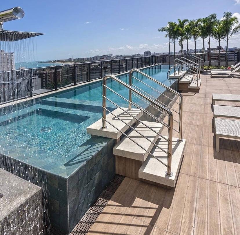An inviting rooftop pool area features a waterfall and steps leading into the water. Surrounding lounge chairs offer a space to relax, while palm trees provide a touch of greenery against a clear blue sky.