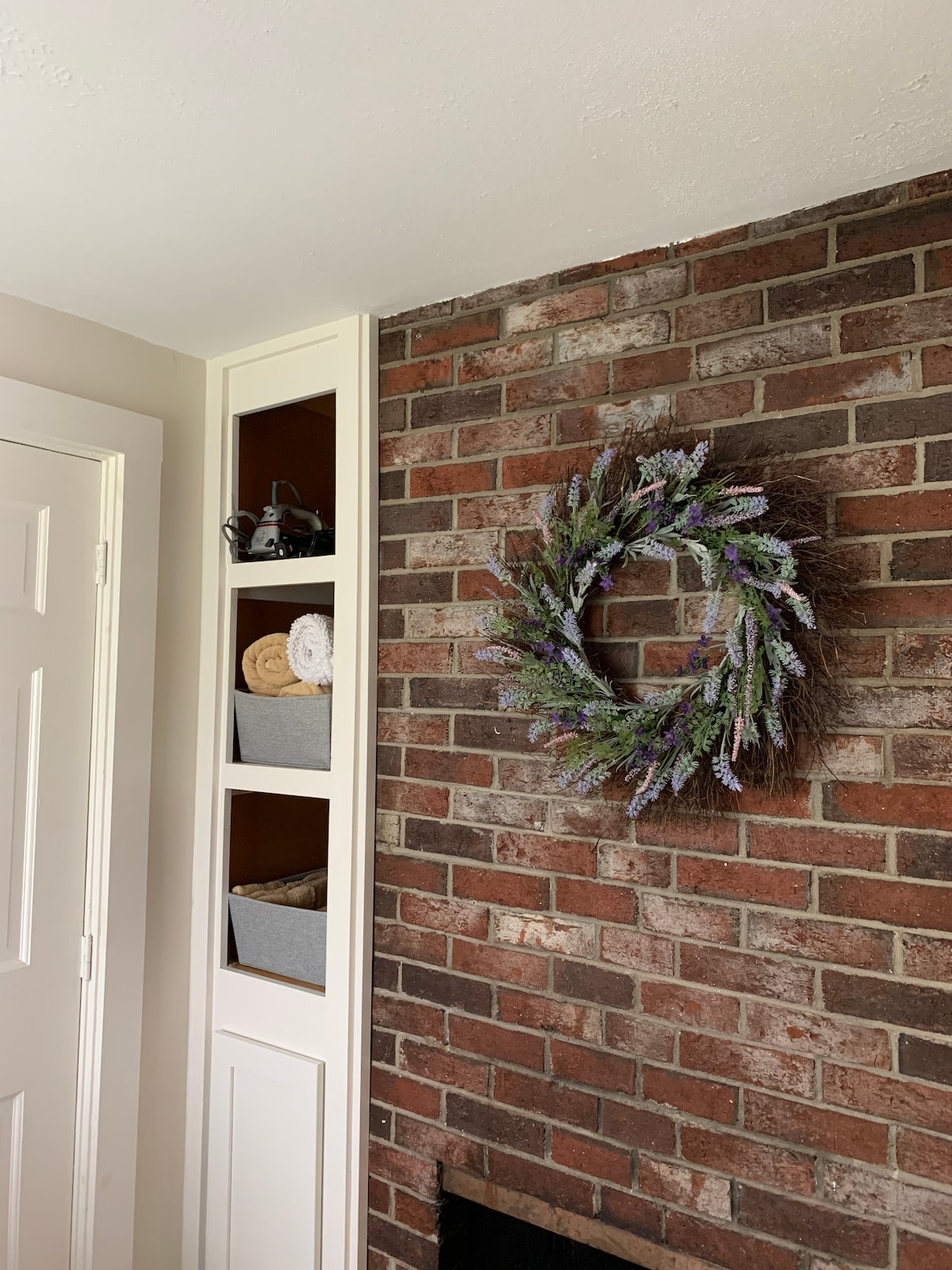 A brick wall is complemented by a decorative wreath of greenery and lavender. Shelving on the adjacent wall contains woven baskets and rolled towels, adding a functional element to the entryway. A white door provides access to the space.