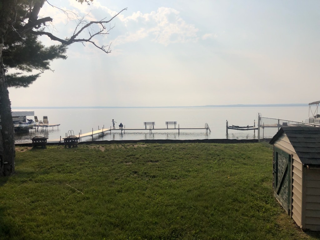 A serene lakefront view features a grassy area leading to a wooden dock extending into calm water. A small shed sits to the right, and distant boats are visible. Soft light filters through the clouds, enhancing the tranquil atmosphere of the setting.