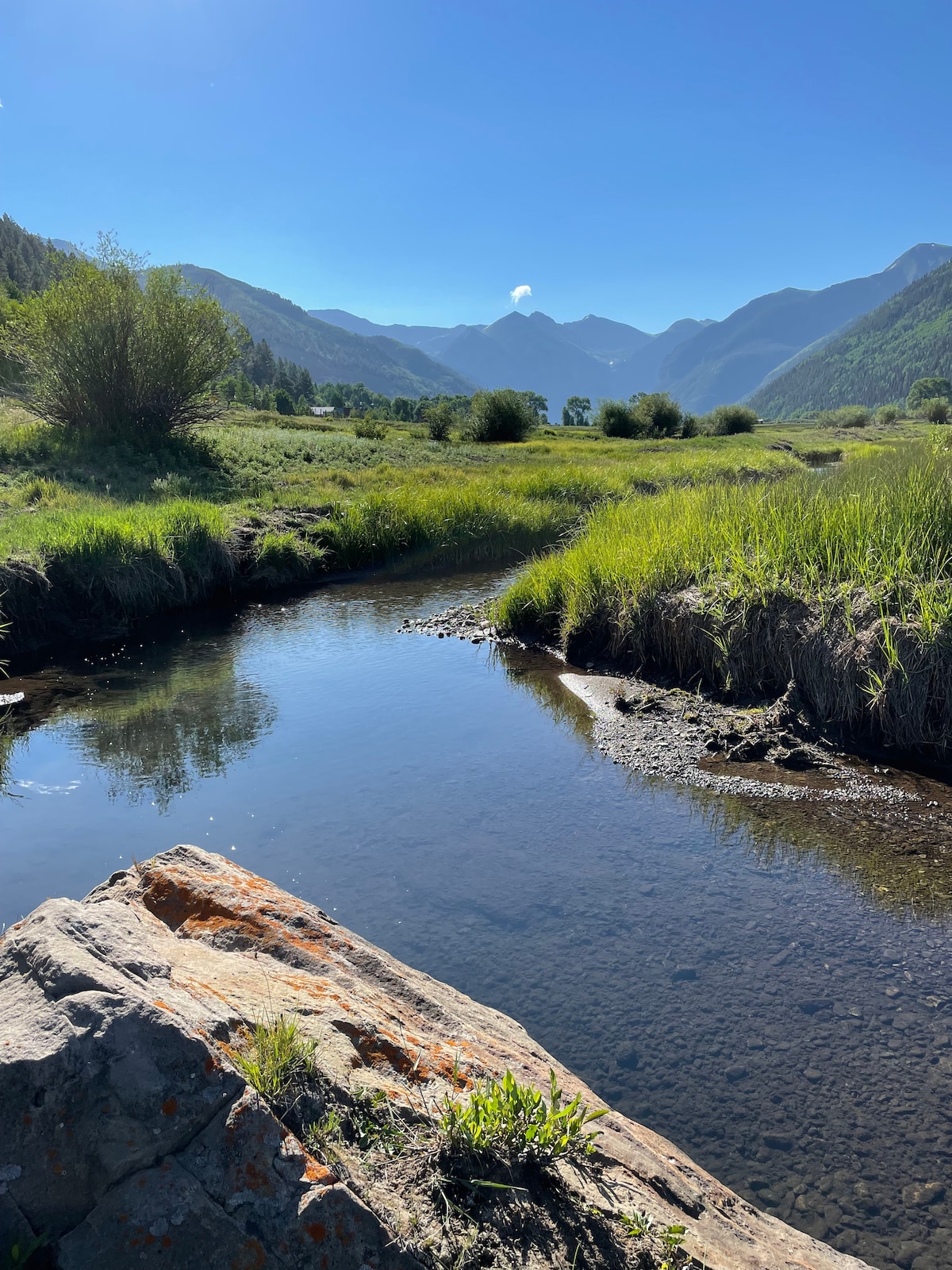 A serene mountain scene features a flowing stream reflecting the clear blue sky. Lush greenery borders the water, and distant mountains create a picturesque backdrop. An outcropping of rock highlights the natural landscape, suggesting a peaceful environment for outdoor activities.